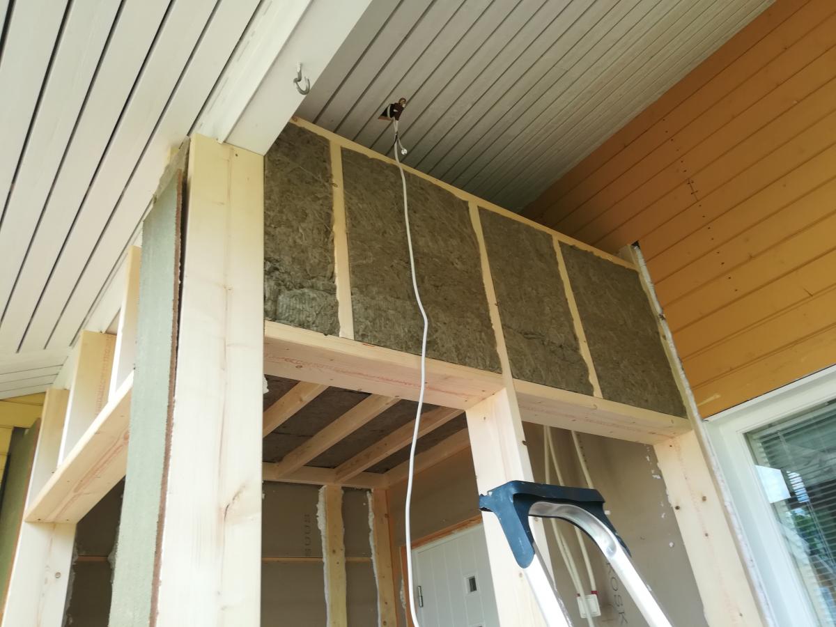 Lähikuva rakenteilla olevasta puurunkoisesta seinästä, jossa on asennettuna harmahtavia mineraalivillaeristeitä. Yläpuolella on valkoisia ja keltaoransseja puupaneelikattoja. Valkoinen sähköjohto roikkuu keskellä. Oikealla alhaalla näkyy osittain ikkuna ja tikkaat. Kohtaus on kirkkaasti valaistu.