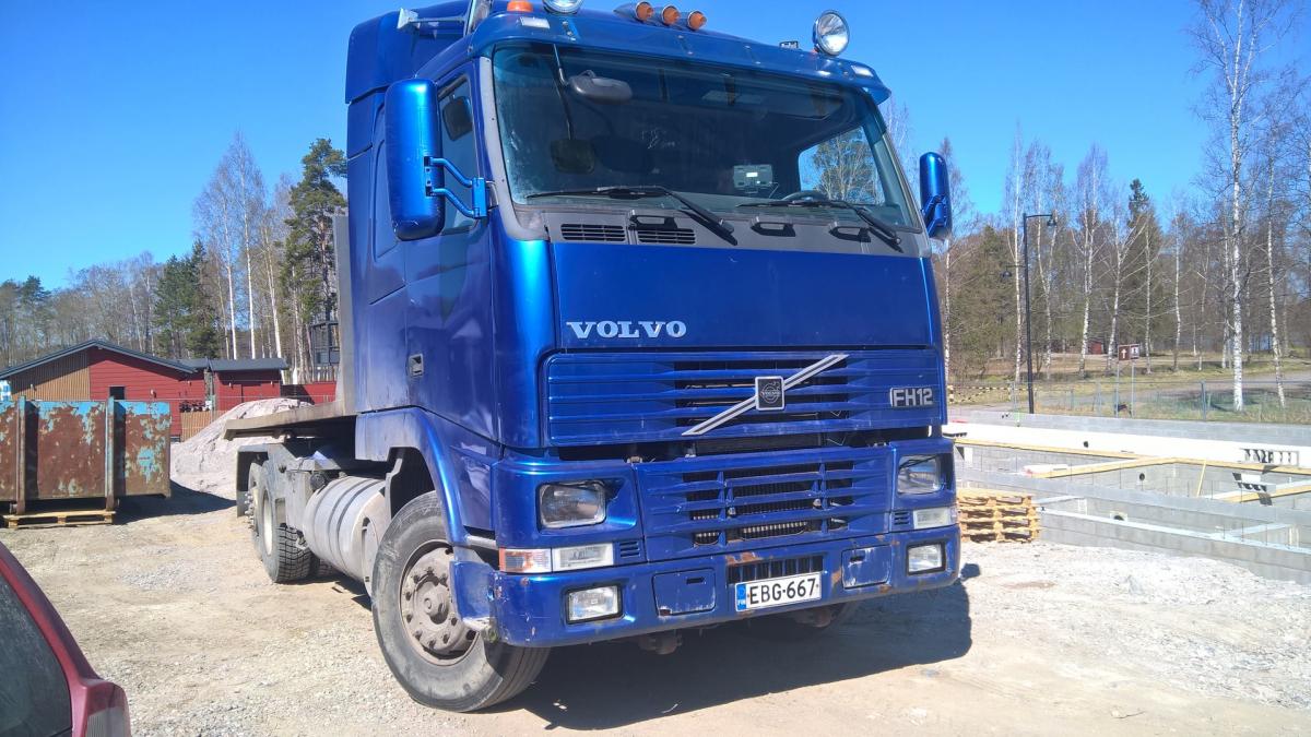 Kirkkaan sininen Volvo FH12 -kuorma-auto on pysäköity sorapihalle aurinkoisena päivänä. Taustalla näkyy punaisia rakennuksia, ruosteisia kontteja ja rakennustyömaa betonivaluineen. Taivas on kirkkaan sininen ja puut ovat vielä lehdettömiä.