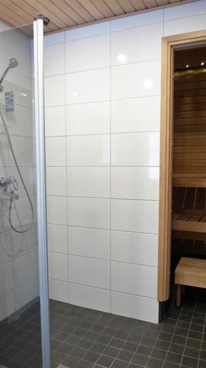 Moderni kylpyhuone, jossa on suihku ja pääsy saunaan. Vasemmalla on suihkutila lasiseinällä, tummanharmaalla laattalattialla ja valkoisilla seinälaatoilla. Keskellä on valkoinen laattaseinä. Oikealla näkyy puinen ovenkarmi ja saunan sisätilat puisine lauteineen. Katto on vaaleaa puupaneelia.
