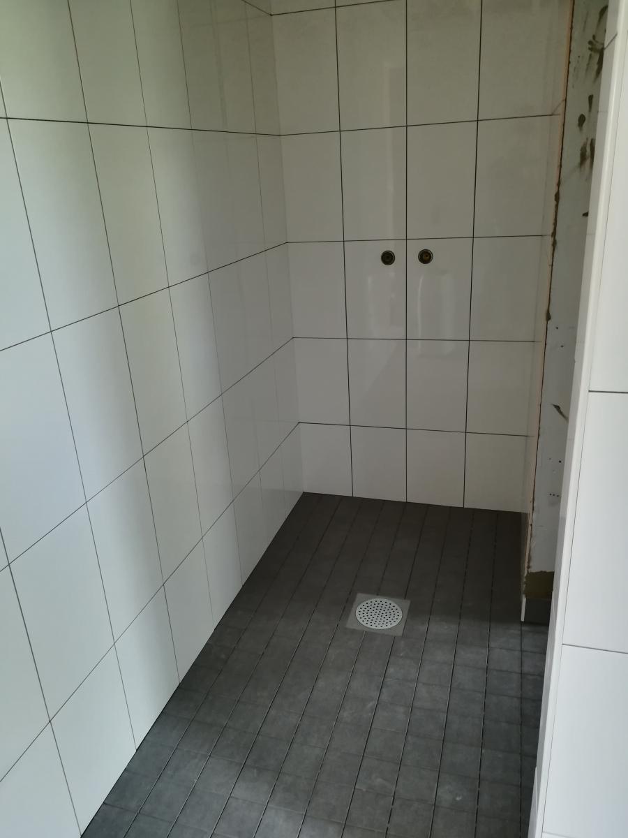 Valkoisilla seinälaatoilla ja tummanharmailla lattialaatoilla laatoitettu suihkutila. Lattialla on pyöreä lattiakaivo. Takaseinässä on kaksi reikää suihkuhanoja varten. Oikea seinä on osittain keskeneräinen.