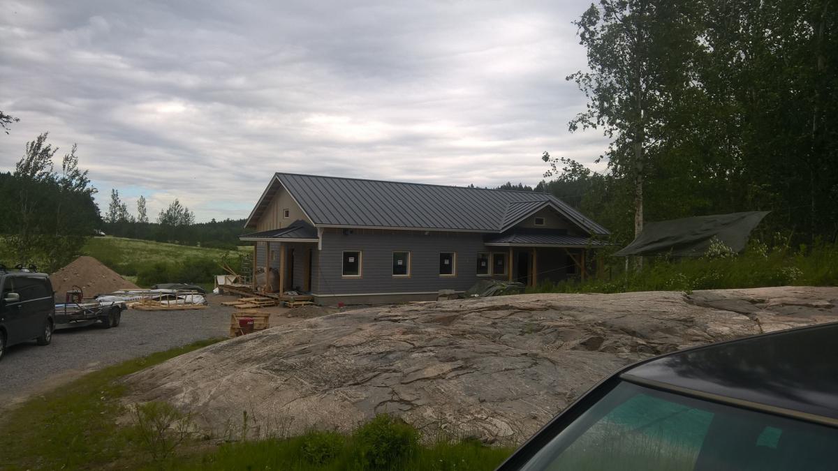 Rakenteilla oleva harmaa talo tummanharmaalla peltikatolla ja useilla ikkunoilla. Etualalla on suuri, vaaleanharmaa kallio ja oikealla näkyy osittain mustan auton katto ja tuulilasi. Vasemmalla on soratie, tumma pakettiauto peräkärryineen, hiekkakasa ja rakennustarvikkeita. Taustalla näkyy vihreä pelto, puita ja metsää. Oikealla talon takana on pressulla peitetty vaja. Taivas on pilvinen ja valaistus tasainen.