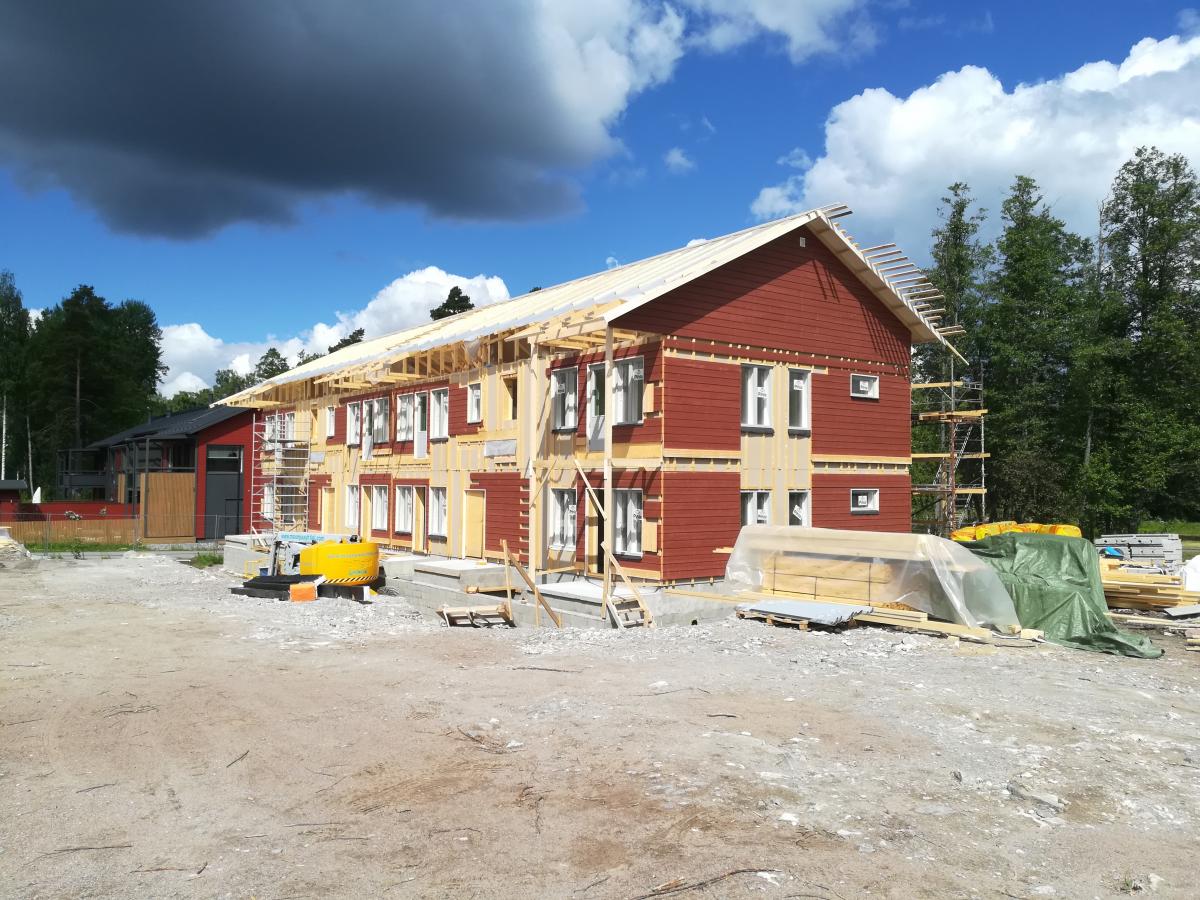 Kuva rakenteilla olevasta kaksikerroksisesta talosta aurinkoisena päivänä. Talossa on punainen puuverhous ja useita ikkunoita, ja sen katto on vielä puurakenteilla. Osa seinistä on vaalean keltaisen eristeen peitossa. Oikealla on rakennustelineitä. Edustalla on soraista maata, keltainen rakennuskone ja pinottuja rakennusmateriaaleja, kuten puutavaraa, peitettynä muovilla ja vihreillä pressuilla. Taustalla on vihreitä puita ja vasemmalla tummempi rakennus. Taivaalla on sinistä, valkoisia pilviä ja vasemmalla tummanharmaita pilviä.