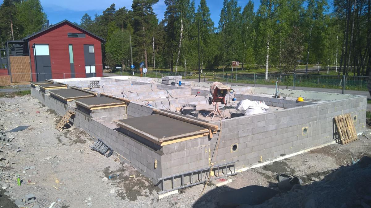 Rakennustyömaa aurinkoisena päivänä. Kuvan etualalla on harmaista betoniharkoista rakennettu talon perustukset, joissa on useita vastavalettuja, puukehyksillä varustettuja aukkoja ja pyöreitä reikiä. Oikealla näkyy sementtimylly ja rakennustarvikkeita. Taustalla vasemmalla on moderni punainen talo ja oikealla metsää sekä tie.