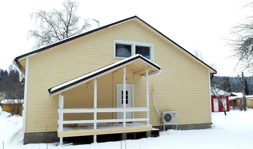 Vaaleankeltainen puutalo talvisessa maisemassa. Talon katolla ja valkoisella kuistilla on lunta ja jääpuikkoja. Kuistilla on valkoiset kaiteet ja valkoinen ovi. Yläkerran ikkunat ovat huurussa. Talon oikealla puolella on ilmastointilaitteen ulkoyksikkö. Taustalla näkyy lumisia puita ja muita rakennuksia pilvisenä päivänä.
