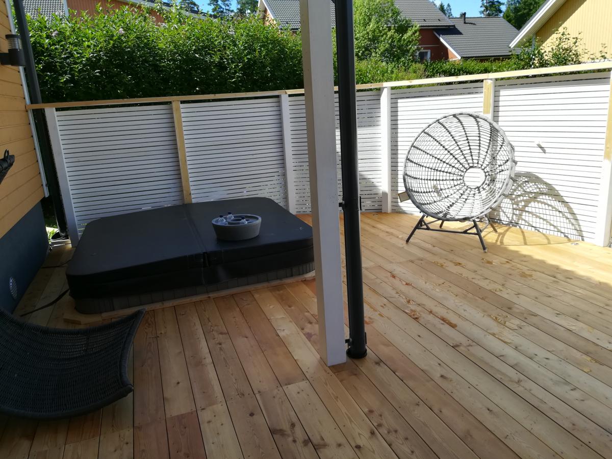 Aurinkoinen kuva puuterassista, jossa on tummanharmaa peitetty poreallas ja musta rottinkinen pyöreä tuoli. Terassin takana on valkoinen säleaita ja vihreitä pensaita. Tuoli heittää pitkän varjon vaaleanruskealle puulattialle. Vasemmassa alakulmassa näkyy osittain toinen tumma tuoli.