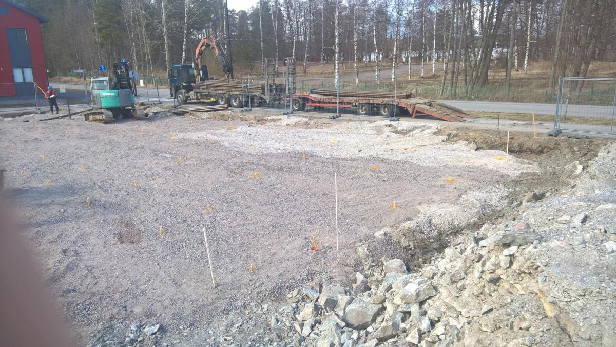 Kuva rakennustyömaasta kirkkaassa päivänvalossa. Etualalla on vaaleanharmaata soraa ja maata, jossa on useita keltaisia merkkejä ja muutama valkoinen keppi. Oikealla on kasa kiviä ja kaivettua maata. Keskellä näkyy tie, jonka varrella on nosturilla varustettu kuorma-auto ja lavettiperävaunu, molemmat lastattuina pitkillä metalliputkilla. Vasemmalla on vihreä kaivinkone ja punatakkinen henkilö pitelemässä keltaista tankoa. Taustalla on puita ja punainen rakennus.