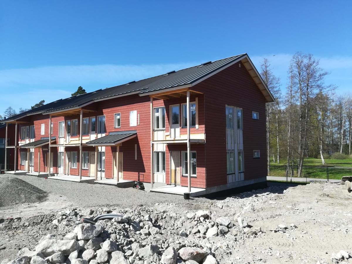Kuva esittää rivin punaruskeita, kaksikerroksisia rivitaloja tai asuntoja rakennusvaiheessa kirkkaan sinisen taivaan alla. Rakennuksissa on tummat katot, katetut sisäänkäynnit ja toisen kerroksen parvekkeet. Etualalla on rakennustyömaa soraa ja kiviä, ja taustalla näkyy puita ja vihreää aluetta.