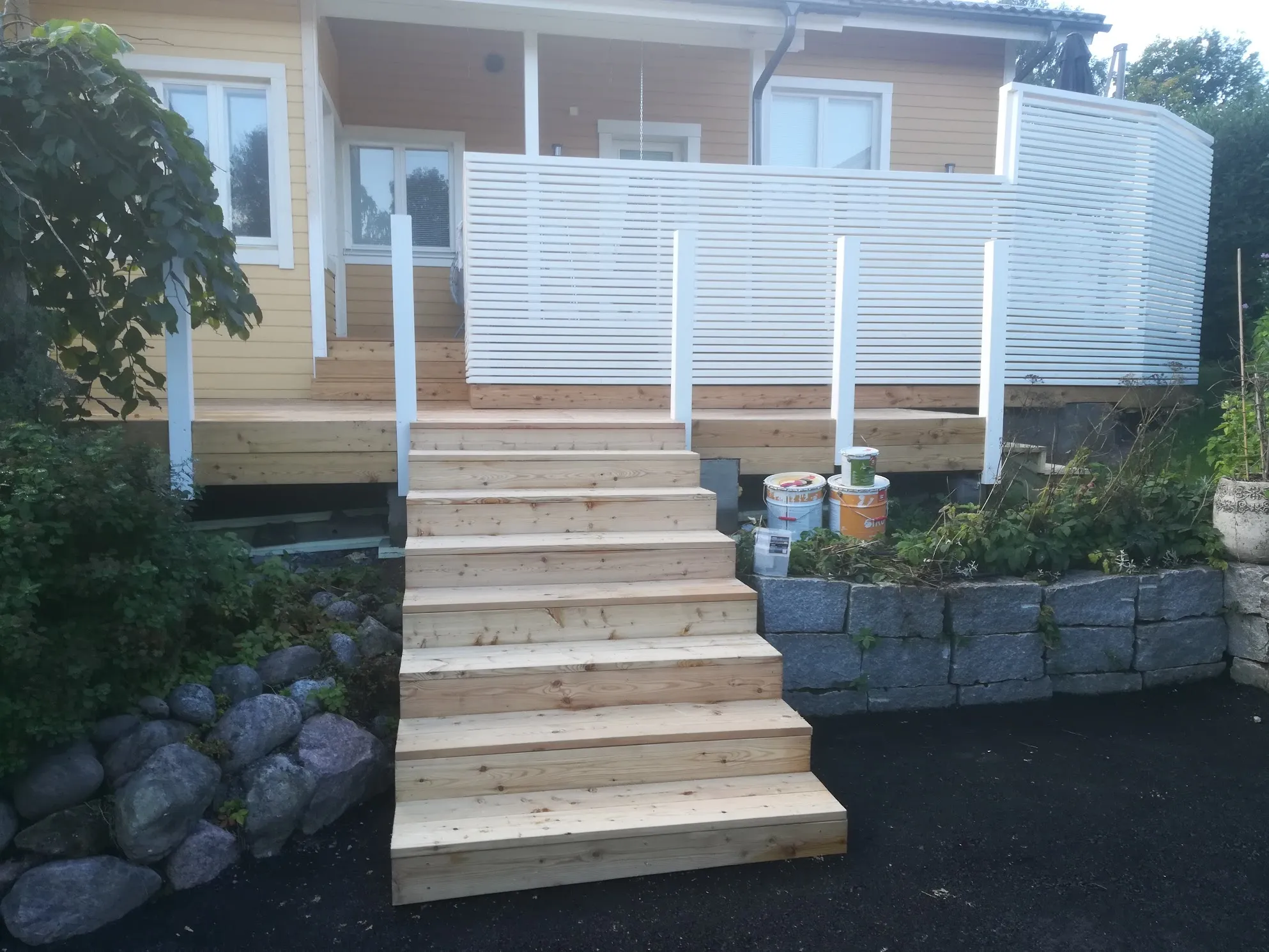 Keltaisen puutalon edessä on uusi, vaaleasta puusta rakennettu terassi ja portaat. Terassilla on valkoinen vaakalaudoitettu näkösuoja. Vasemmalla on vihreää kasvillisuutta ja harmaita kiviä. Oikealla näkyy harmaa kivimuuri ja muutama maalipurkki. Etualalla on tumma piha-alue kirkkaassa päivänvalossa.